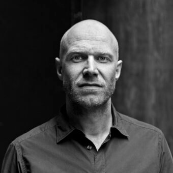 Black and white portrait of Maarten de Ceulaer, a man with a bald head and grey beard, wearing a dark button-down shirt against a blurred background.