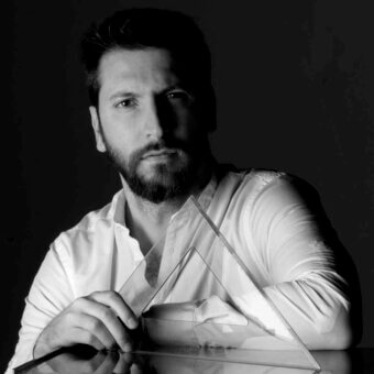 Black and white portrait of Marco Spatti with dark hair, beard, white shirt, and triangular prism, studio lighting.