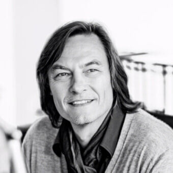 Black and white portrait of Michael Schmidt, a man with long dark hair, wearing a gray sweater and collared shirt, gentle smile.