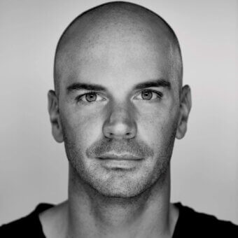 Portrait of Benjamin Hubert, a man with shaved head and mustache, against light gray, displaying natural skin texture and direct gaze.