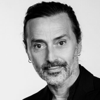 Fabio Novembre portrait, black and white photograph, man with beard, silver earrings, dark jacket, neutral background, direct gaze.