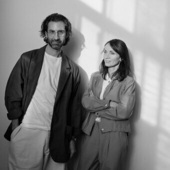 Black and white portrait of two designers, man in blazer and trousers, woman in grey jumpsuit, against light grey wall with diagonal shadows.