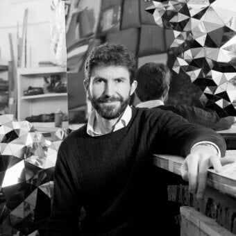 Black and white portrait of Jake Phipps leaning against wood, overlaid with grayscale geometric triangles, in a workshop setting.