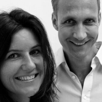 Black and white portrait of a smiling woman and man, both with fair skin, facing forward against a light gray backdrop.