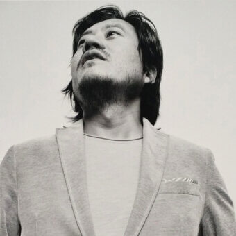 Black and white portrait of Sean Yoo wearing a light gray textured blazer, white t-shirt, and black choker, with long dark hair and beard.