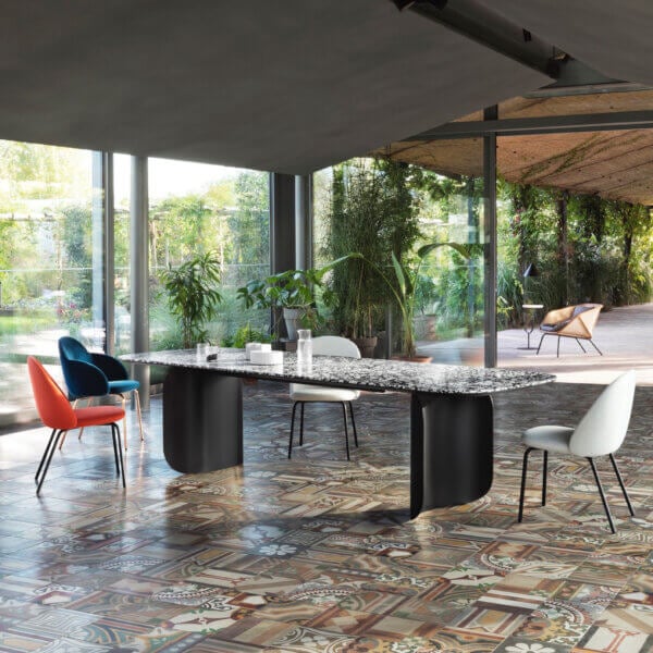 Dining table with dark grey and white mosaic top, surrounded by orange and white chairs, in a room with patterned terracotta flooring and glass walls.