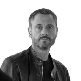 Davide Carlesi portrait, black and white, man with dark hair and beard wearing a black leather jacket, neutral expression, studio lighting.