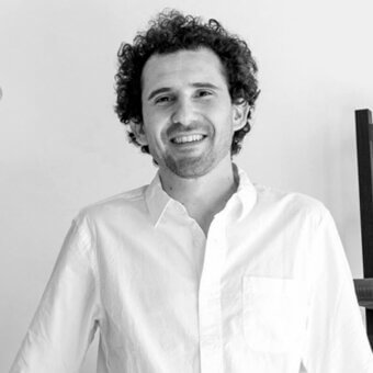 Man portrait, black and white, dark curly hair, short beard, light shirt, plain wall background, soft lighting, relaxed expression.
