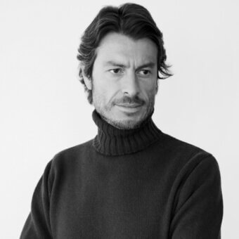 Man portrait, grayscale, dark hair with gray streaks, neutral expression, black turtleneck sweater, plain light gray background, diffused lighting.