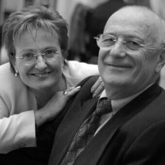 Black and white portrait of a couple, man in suit and tie, woman with layered hair, displaying affection and wearing jewelry.