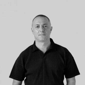 Man portrait, black polo shirt with pleated detailing, shaved head, neutral gray background, diffused studio lighting, direct gaze.