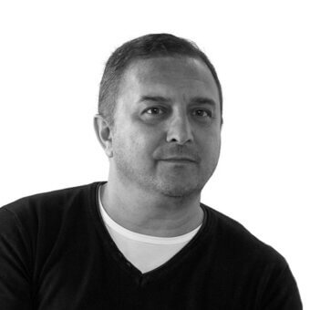 Black and white portrait of Luca Tonelli, short dark hair, black v-neck sweater, neutral expression, head and shoulders composition.