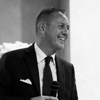 Marco Pozoli portrait, black suit, white shirt, holding black microphone, blurred event background, soft lighting, formal attire.