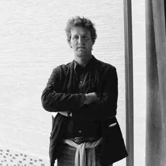 Black and white portrait of Massimiliano Mornati with crossed arms, wearing a jacket and scarf, against blurred window blinds.