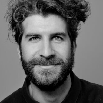 Man portrait, gray scale, dark curly hair and full beard, wearing a dark turtleneck sweater, neutral gray background, subtle expression.