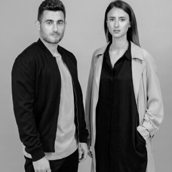 Black and white portrait of a couple, man in bomber jacket and woman in black dress with beige and black coat, gray background.
