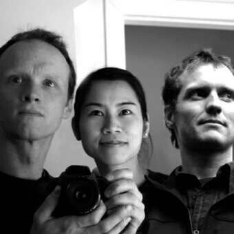 Black and white portrait of three men, central figure holding a digital camera, casual clothing, plain white wall background.
