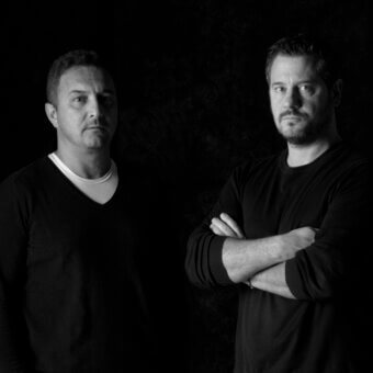 Black and white portrait of two men with neutral expressions against a dark, mottled textured background, wearing black sweaters.