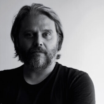 Portrait of Cristian Malisan, gray-toned hair and beard, wearing a dark crew-neck shirt, against a light gray backdrop, direct gaze.
