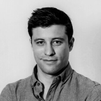 Black and white portrait of a man with short dark hair, fair skin, and a light button-down shirt, arms crossed, against a neutral wall.