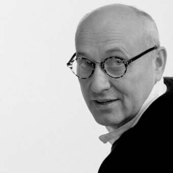 Black and white portrait of Ennio Arosio, a man in a dark suit, wire glasses, and bow tie, facing right against a light gray backdrop.