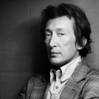 Black and white portrait of Guglielmo Berchicci wearing a plaid blazer with an open-collar white shirt, against a light paneled wall.