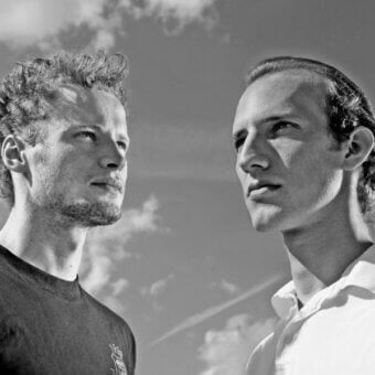 Black and white portrait of two men with contrasting hair, looking upwards against a cloudy sky backdrop, studio lighting.
