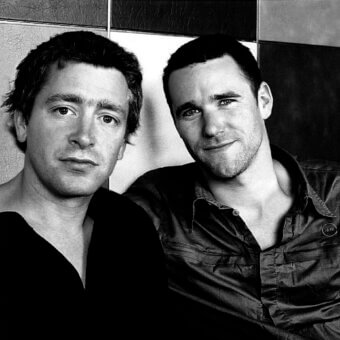 Black and white portrait of two men against tiled backdrop, featuring textured surfaces, contrasting shadows, and casual attire.