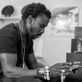 Ini Archibong portrait, black and white, seated man with dreadlocks holding a smartphone, wearing black shirt and silver jewelry, blurred interior background.
