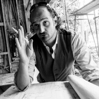 Marcello Pozzi portrait, blueprints, construction site, tweed vest, sunglasses, pencil, technical drawings, exposed wood framing, diffused lighting.
