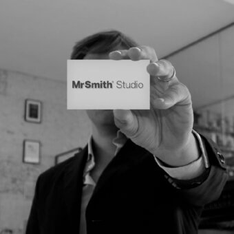 Person holding business card reading "Mr Smith' Studio" wearing a suit, blurred studio background with brick wall and framed artwork.