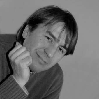 Black and white portrait of Nicola Nerboni, a man with dark hair wearing a brown ribbed knit sweater, hand to chin, neutral gray background.