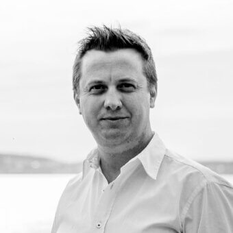 Black and white portrait of Noé Duchaufour Lawrance, fair complexion, short dark hair, relaxed expression, light shirt, blurred outdoor background.