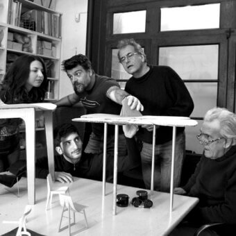 Design studio scene with five people discussing miniature chair models on a table, black and white, bookshelf backdrop.
