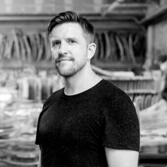 Black and white portrait of Samuel Wilkinson wearing a textured black t-shirt, in a workshop with stacked wood forms.