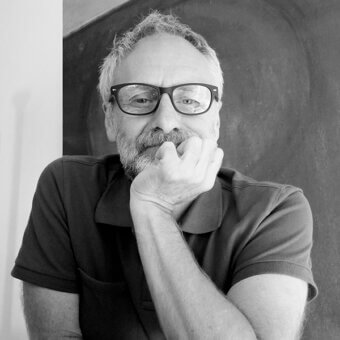 Black and white portrait of Sergio Brioschi, a man with gray hair and beard, wearing glasses and a dark polo shirt, hands clasped in thought.