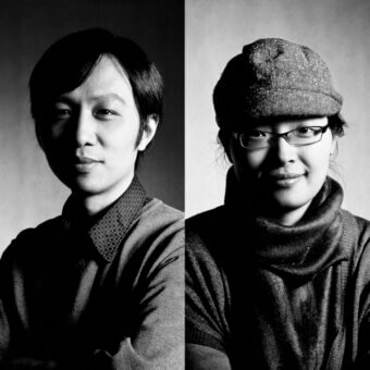 Black and white studio portrait of two people with crossed arms, wearing gray sweaters and knit cap, against a light gray backdrop.