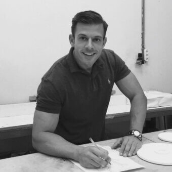 Chris Basias sketches at a workbench, wearing a Ralph Lauren polo, with a watch and pencil visible, amidst papers and ceramic plates.