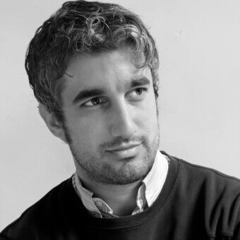 Black and white portrait of Jordi Pla, a man with dark wavy hair, beard, and neutral expression wearing a dark sweater and white collared shirt.