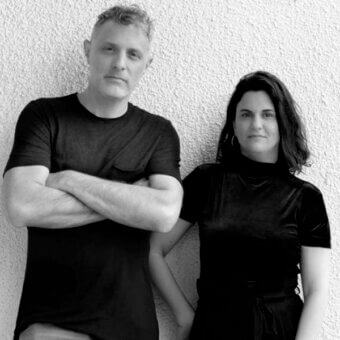 Black and white portrait of a man in a t-shirt and a woman in a black velvet top and skirt, against a textured off-white stucco wall.