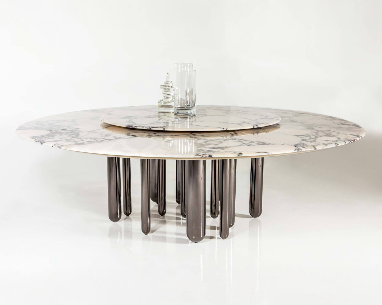 Carrara marble dining table with dark gray metal legs and brass rotating base, featuring gray veining and clear glass vases.