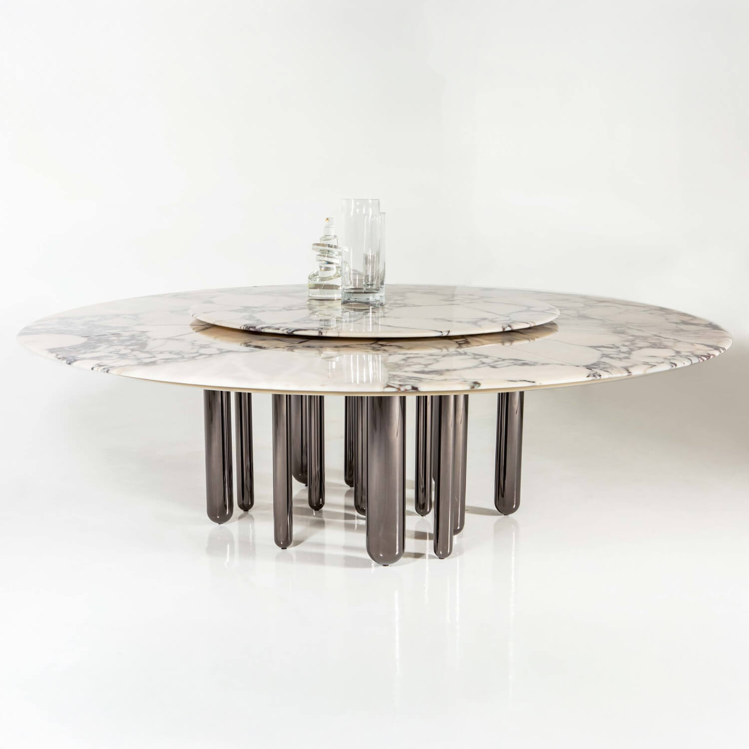 Marble dining table with gray veining, eleven dark gray cylindrical legs, and clear glass vases centered on polished white floor.