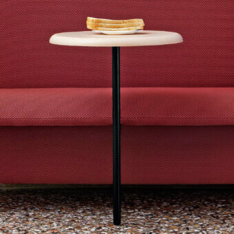 Circular coffee table with light wood top and black metal leg, set before a red textured banquette on terrazzo flooring.