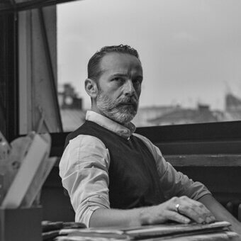 Black and white portrait of Emanuele Genuizzi seated at a dark wood desk, wearing a white shirt and knit vest, with a cityscape visible through the window.