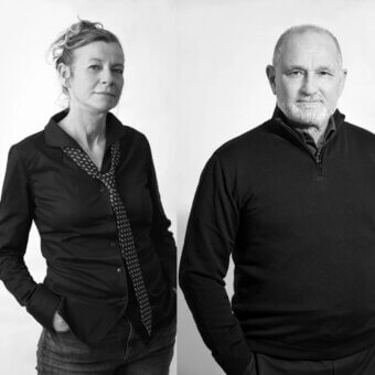 Black and white portrait of two people, a woman in a black shirt and scarf, and a man in a gray sweater, against a white backdrop.