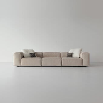Modular sofa, beige leather, rectangular sections with wide armrests and geometric pillows, against a light gray wall.
