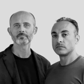 Black and white portrait of two men, one bald with a beard in a blazer, the other with short hair in a t-shirt, against a white backdrop.