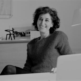 Black and white portrait of Patrizia Bertolini seated with laptop, wearing a dark wool sweater, in a room with wood floors.