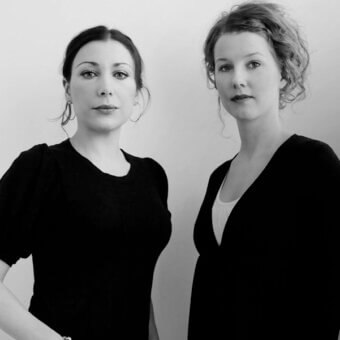 Black and white studio portrait of two women in black dresses, one with a bun and hoops, the other with wavy reddish-brown hair.