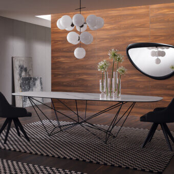 White marble dining table with black geometric metal base, oval shape, paired with black chairs and checkered rug in wood-paneled room.